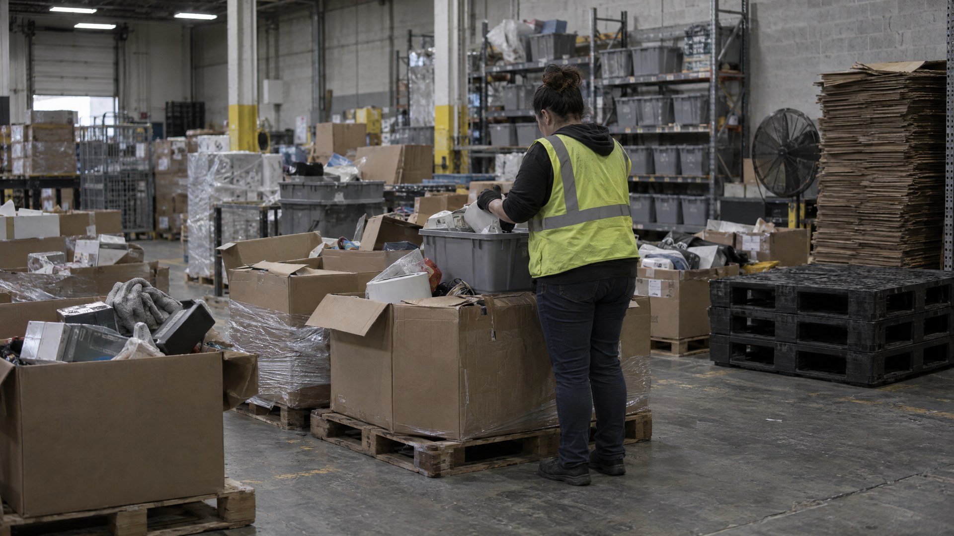 Illustrative returns processing area with pallets of opened cartons and a worker sorting items into a tote.